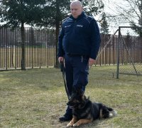 Policjant w umundurowaniu służbowym stoi na podwórku, a przy jego lewej nodze leży pies.