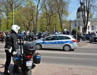Na pierwszym planie policjant przy motocyklu służbowym, w głębi- radiowóz i grupa motocyklistów na tle parku
