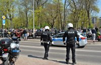 Na pierwszym planie dwóch policjantów w kombinezonach i kaskach, obok nich motocykl policyjny, w głębi grupa motocyklistów
