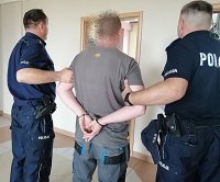 Dwóch policjantów w umundurowaniu służbowym doprowadza pod drzwi mężczyznę w kajdankach na rękach.