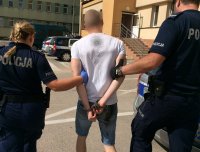 Policjanci w umundurowaniu służbowym prowadzą mężczyznę w białej koszulce z krótkim rękawem, który ma zapięte z tyłu kajdanki na rękach