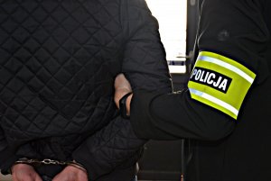 Policjant po cywilnemu z opaska na ramieniu z napisem POLICJA doprowadza zatrzymanego, obaj ubrani na ciemno